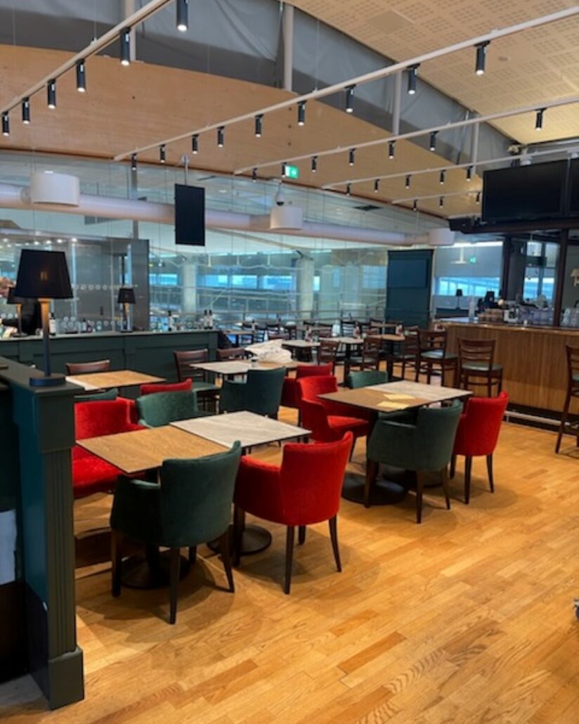 Spiseområde på O'Learys Oslo Lufthavn Gardermoen med trebord, røde og grønne stoler samt baren i bakgrunnen.