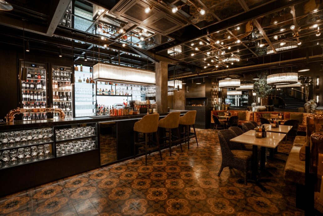 Barområde på Olivia Kluuvi i Finland med flislagt gulv, høye barkrakker, godt opplyst bardisk med flasker og glass, samt stemningsfull restaurantbelysning.