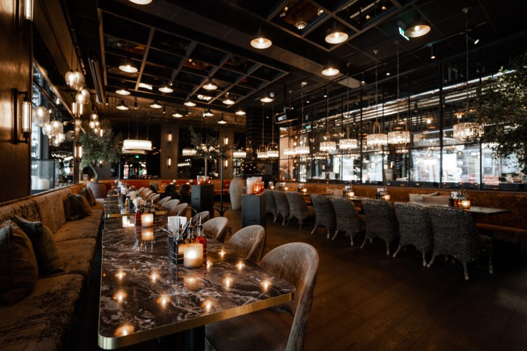 Stemningsfull restaurant på Olivia Lillestrøm med lange marmorbord, dempet belysning, polstrede stoler og speilvegger som reflekterer lysekroner og levende lys.