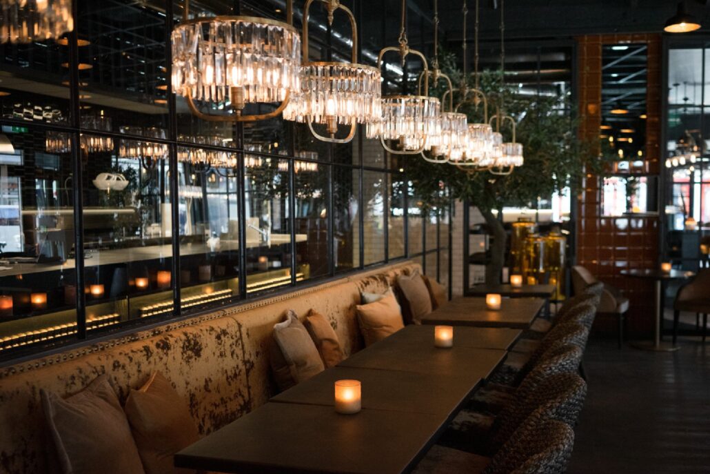Moderne restaurantinteriør på Olivia Lillestrøm med dempet belysning, lange bord med tente stearinlys, polstrede stoler og elegante lysekroner i glass.